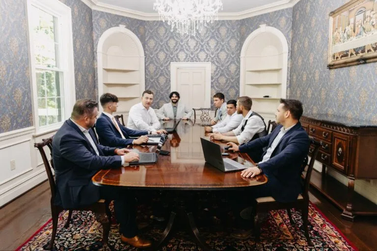 Nine men in business attire sit around a conference table in an elegant room with blue wallpaper, a chandelier, and laptops open for a meeting.